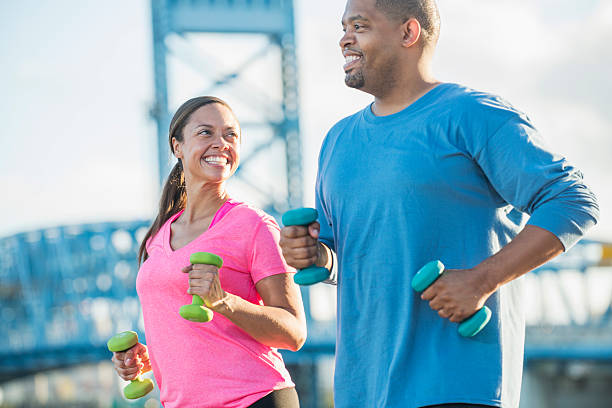 A black couple outdoors on the waterfront of a city, exercising together. They are carrying handweights, power walking, jogging or running on a sunny day to stay in shape.